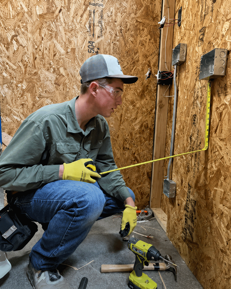 An electrician student in safety glasses measures wiring during a hands-on lab